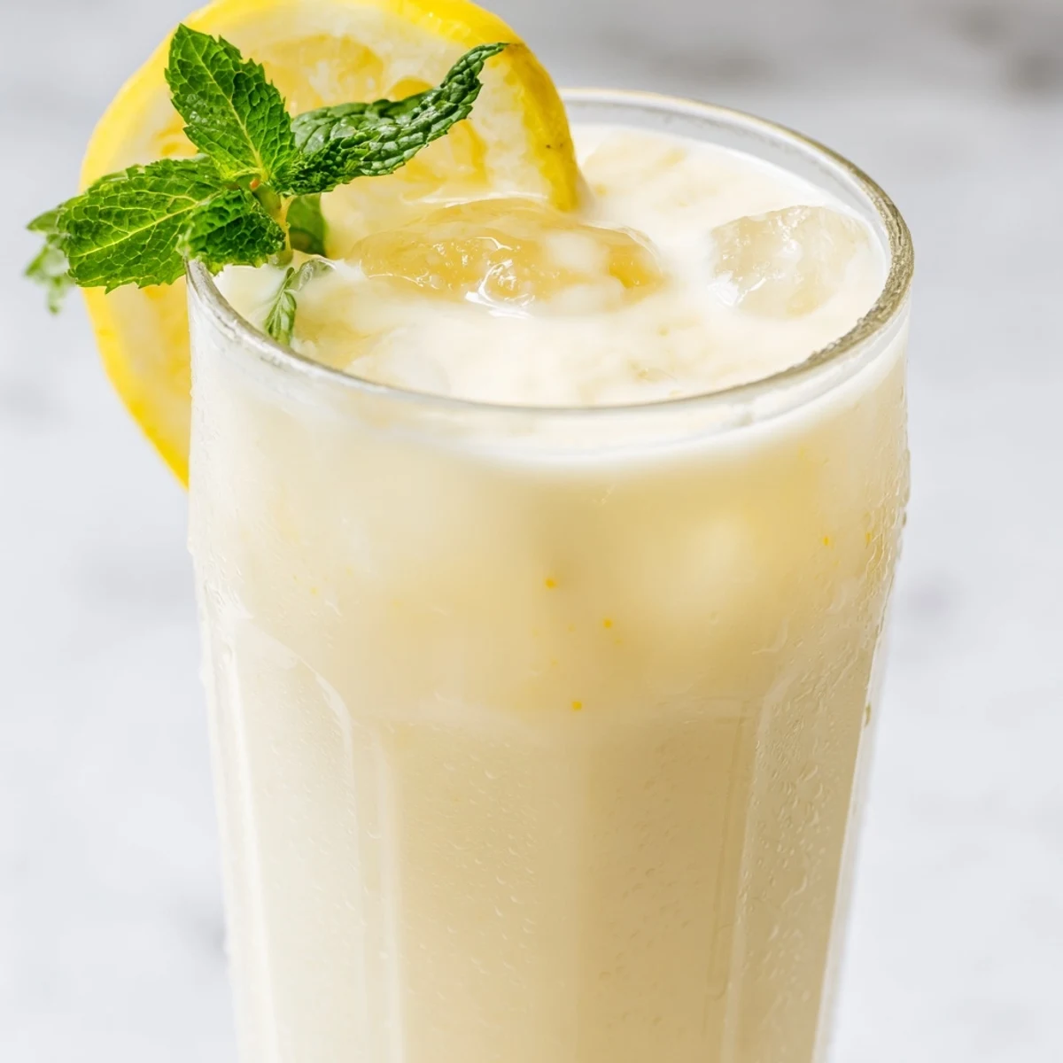 A tall glass of creamy lemonade with ice cubes, lemon slices, and fresh mint leaves on a wooden table.